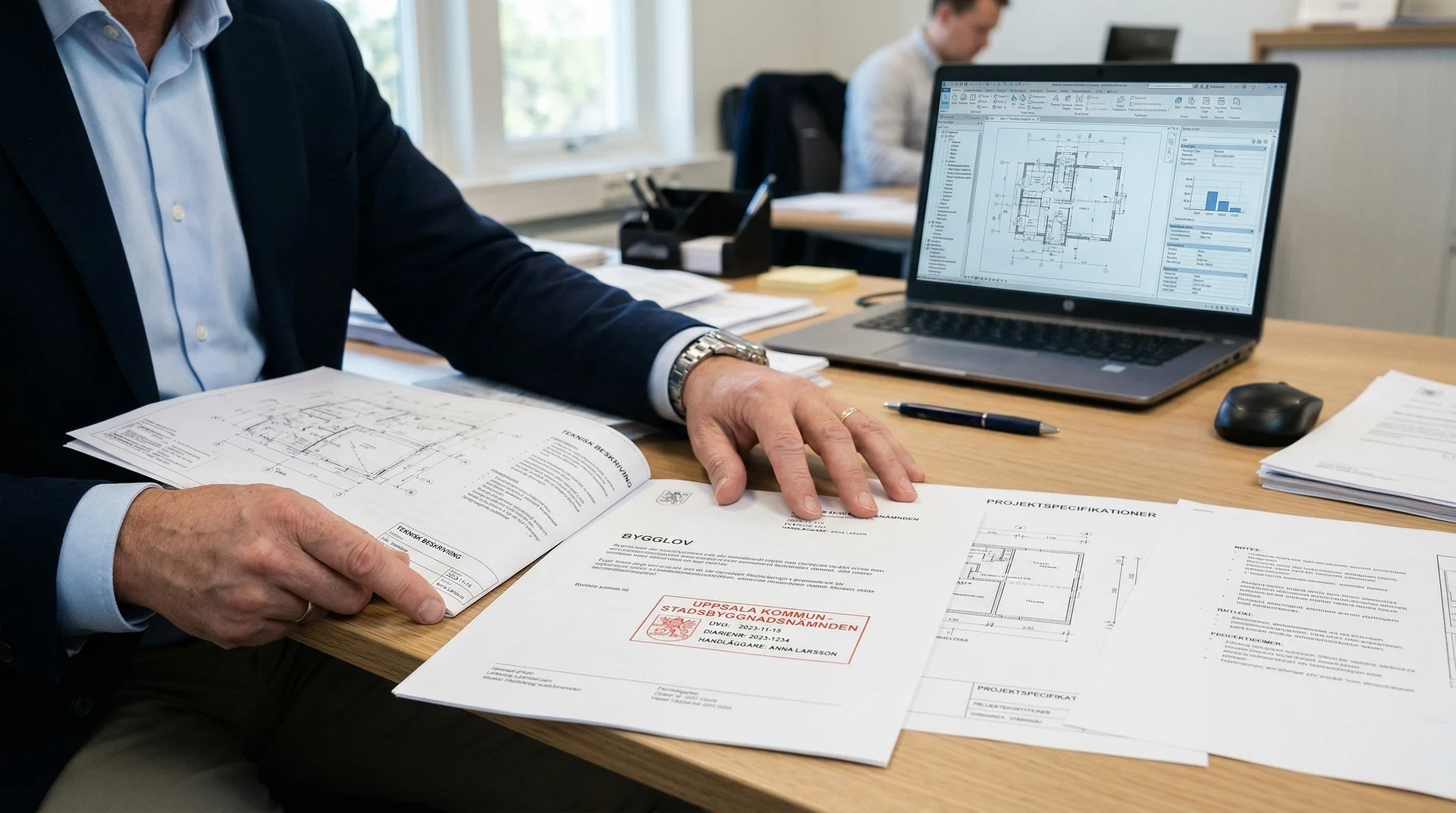 Person i kostym granskar byggritningar och dokument vid skrivbord med laptop.