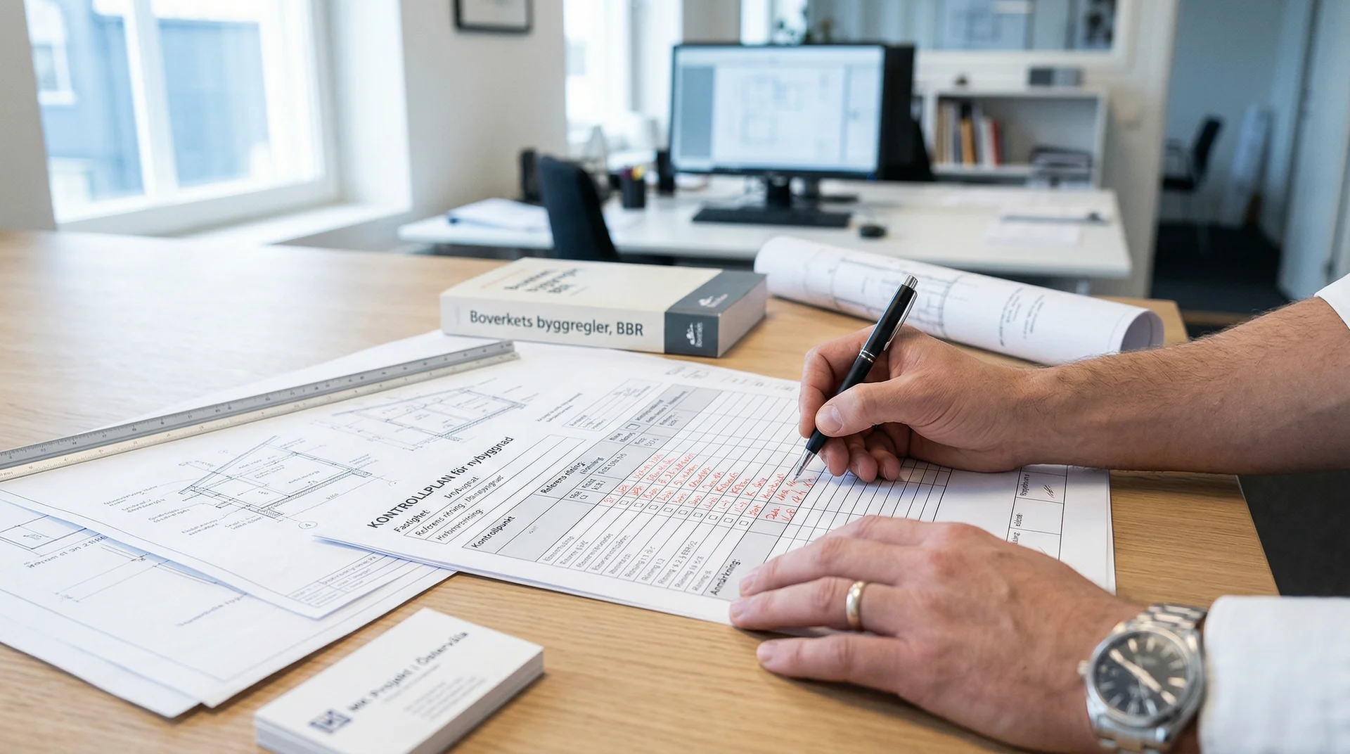 Person fyller i byggdokument vid skrivbord med ritningar och regelbok.